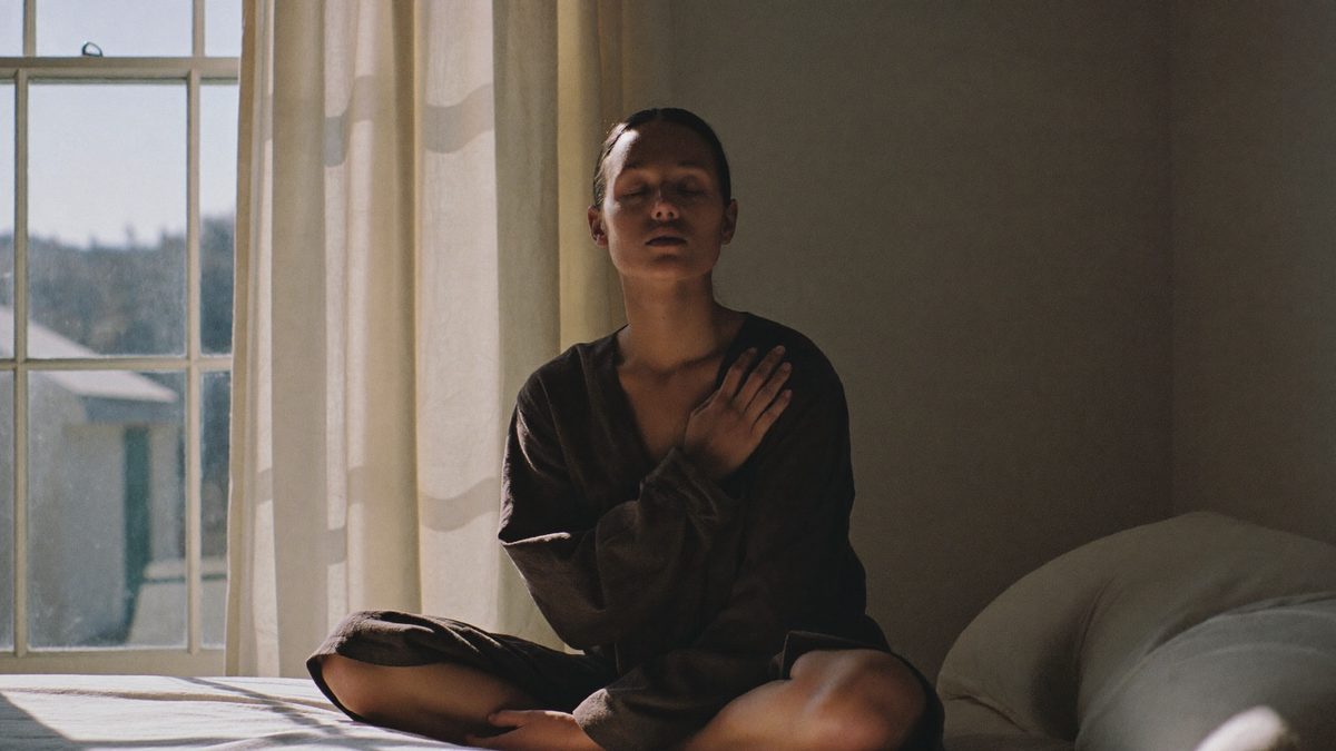 Femme assise en tailleur près d'une fenêtre ensoleillée le matin, yeux fermés, une main sur la poitrine, pratiquant un exercice de respiration calme façon cohérence cardiaque