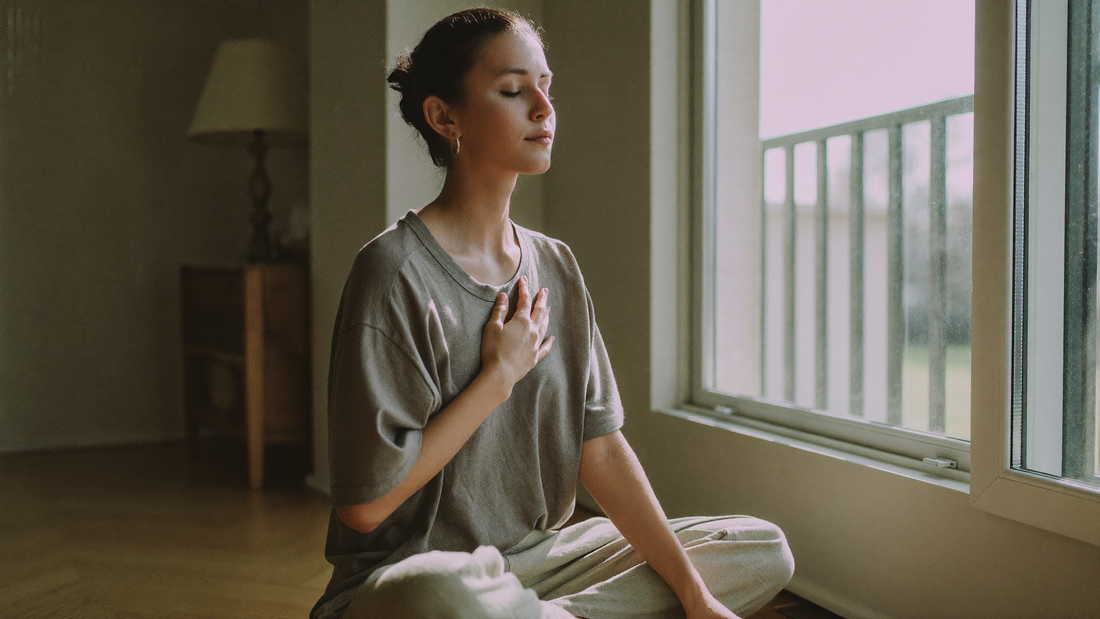 Femme assise en tailleur près d'une fenêtre lumineuse, les yeux fermés, main sur la poitrine, pratiquant un exercice de respiration lente le matin